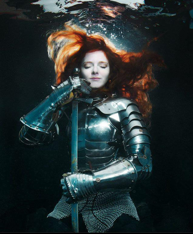 Fotografíad e una mujer con una armadura sumergida en el agua, pelirroja, lleva una espada, ojos cerrados